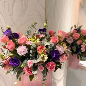 Arreglo floral en base rosa con rosas y flores mixtas en tonos rosa lila y blanco