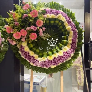 Corona fúnebre rosa y lila de condolencias con flores mixtas, aro blanco y follaje verde en base circular